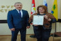 На изображении два человека стоят рядом. Один держит сертификат. На заднем плане видны флаги и надпись на стене.