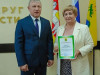 На изображении два человека стоят рядом. Один держит сертификат. На заднем плане видны флаги и надпись на стене.